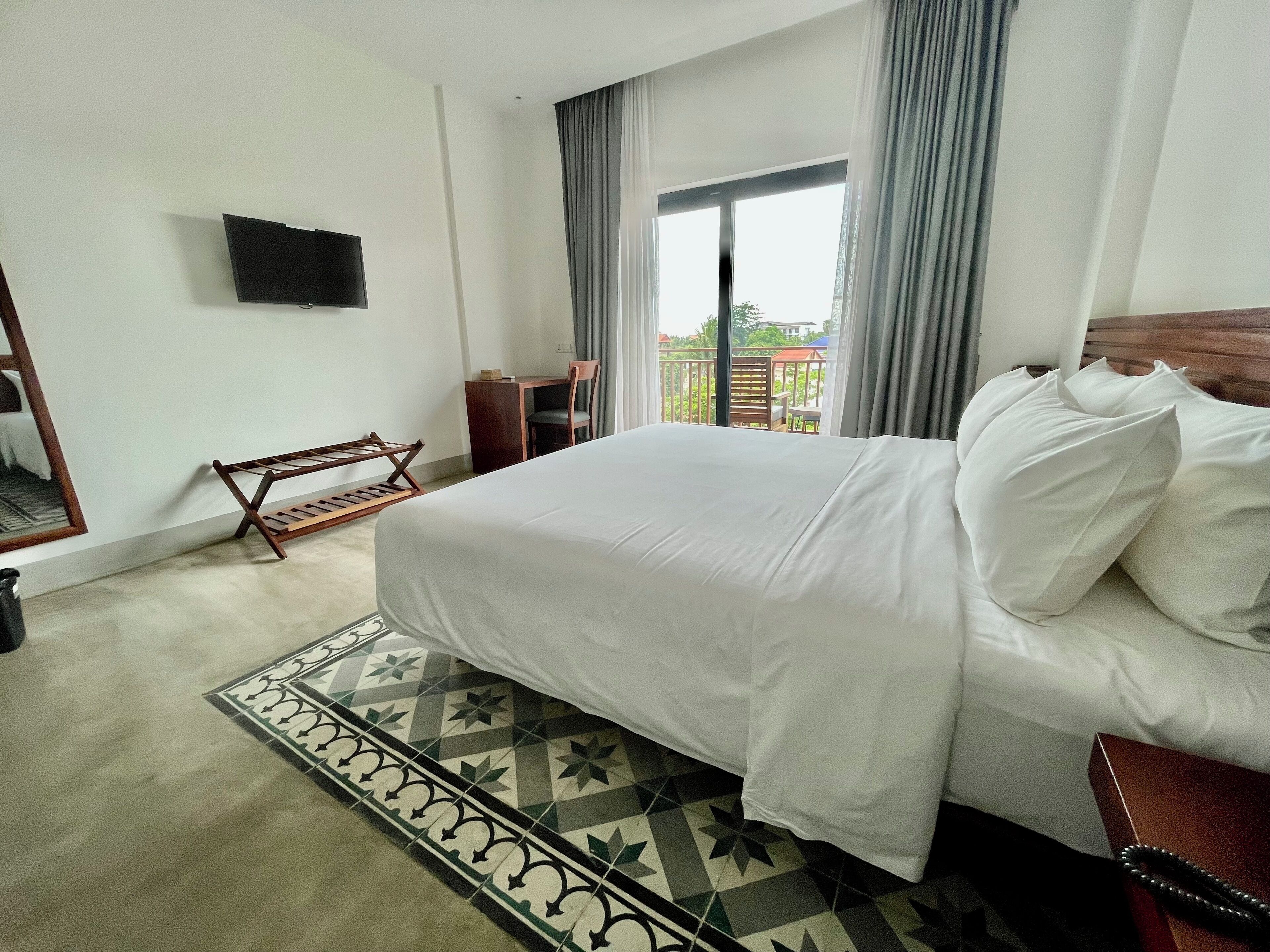A hotel room with a large bed, a desk, a chair, a TV mounted on the wall, and a balcony with a view of the outside.
