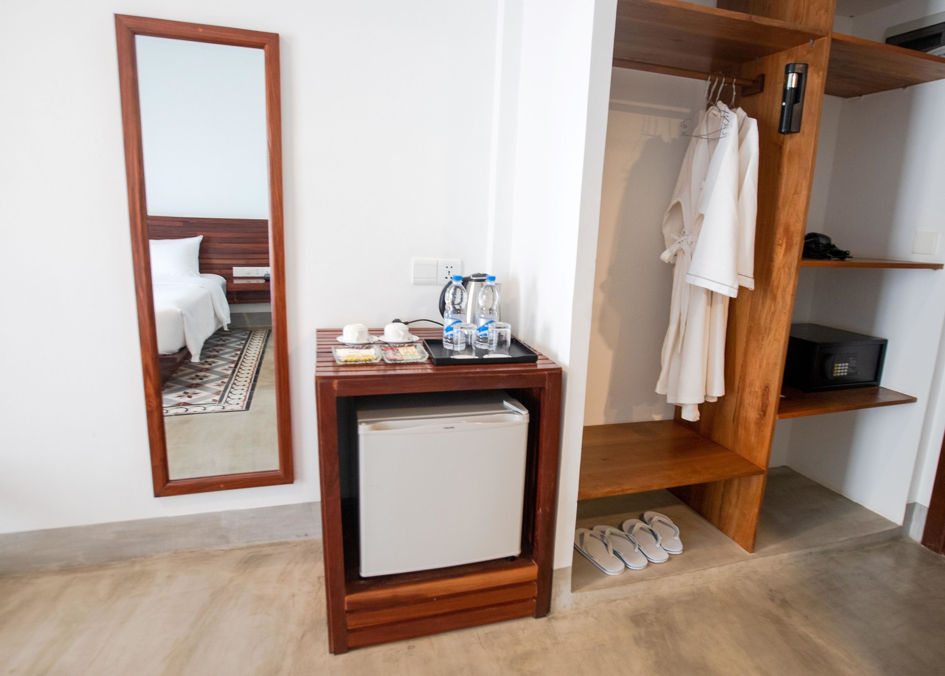 A hotel room with a wooden desk, a small refrigerator, a mirror, a wardrobe, and a pair of slippers on the floor.