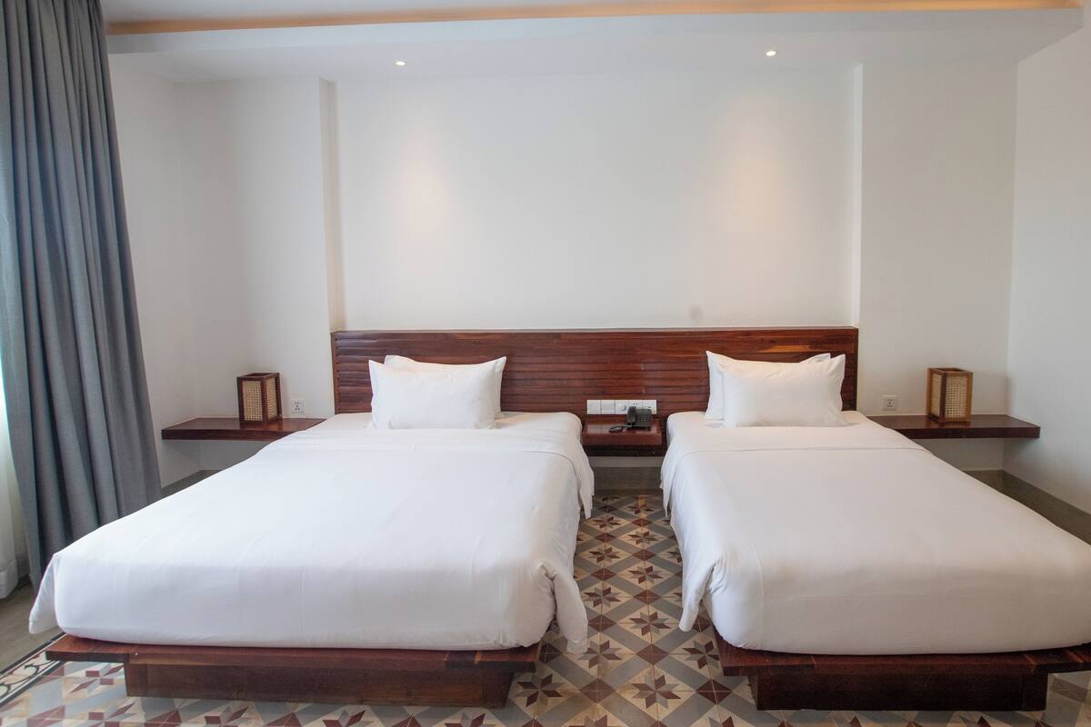 A hotel room with two single beds, wooden headboards, bedside tables with lamps, a patterned floor, and a large window with curtains.