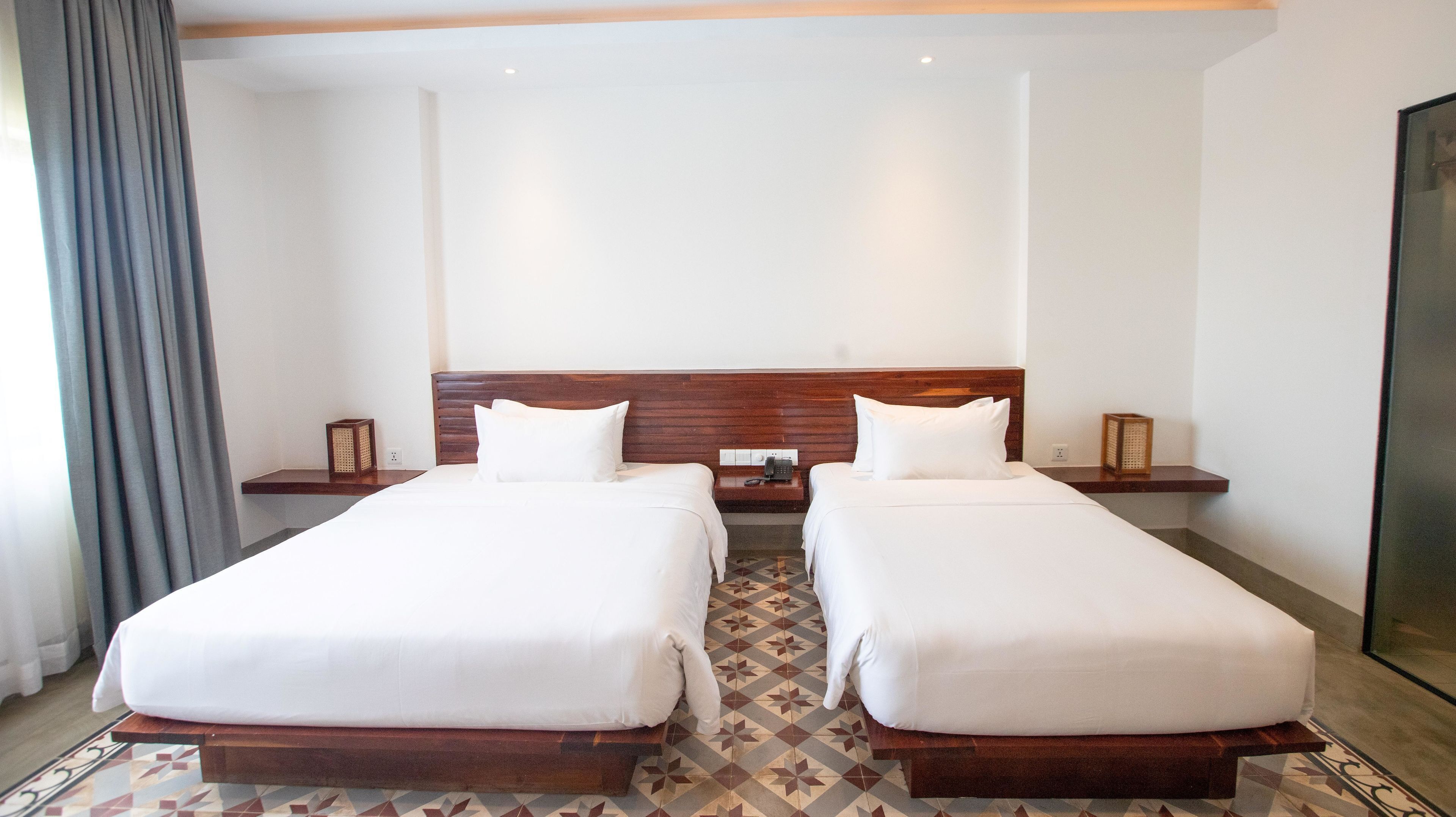 A hotel room with two single beds, wooden headboards, a small bedside table with a lamp, and a patterned floor.