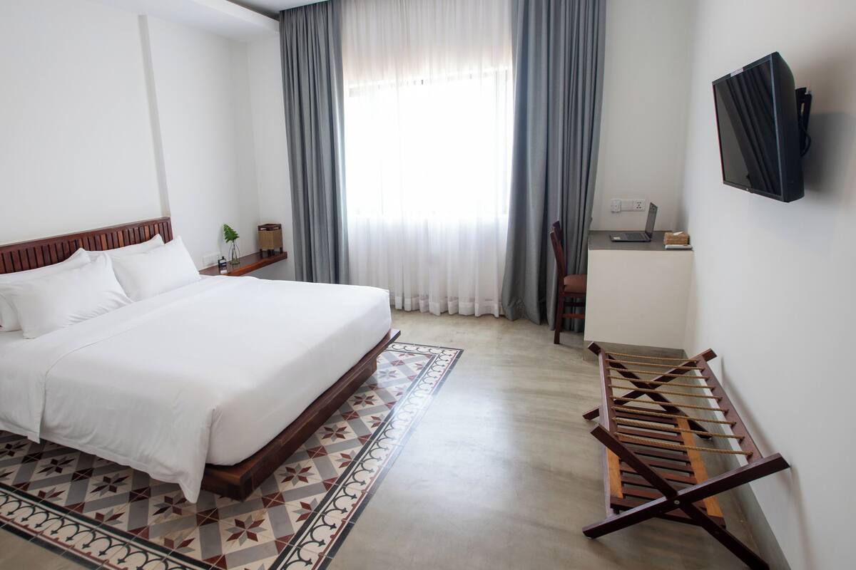 A hotel room with a large bed, a desk with a computer, a TV mounted on the wall, and a window with curtains.