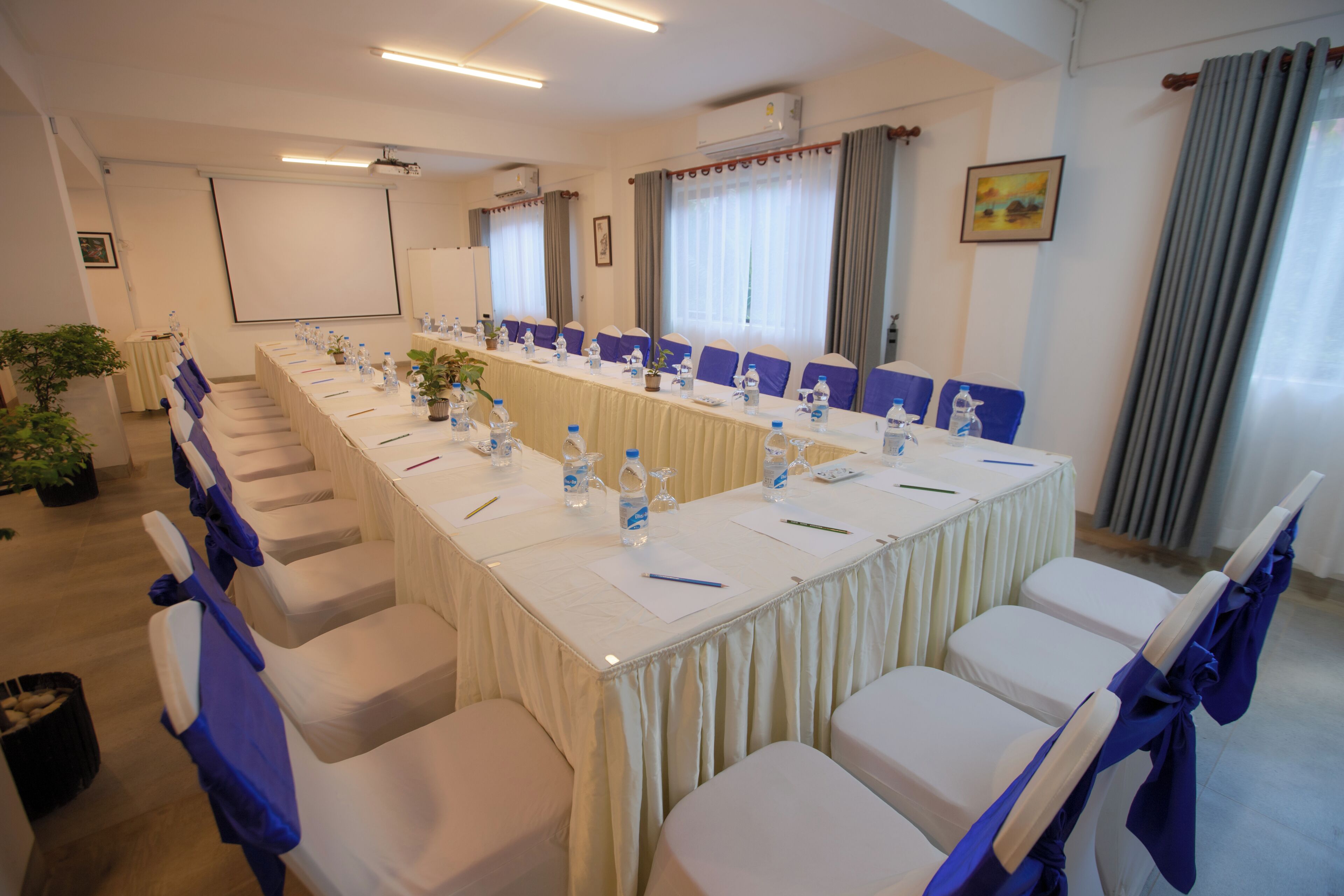 Ein Konferenzraum mit langem Tisch, Stühlen, Wasserflaschen und einer Projektorleinwand.