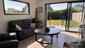 A modern living room with a dark-colored sofa, a glass coffee table, and a view of a garden through large sliding glass doors. - Maison de Vacances à 200 m de L'aber Benoît (Landéda)