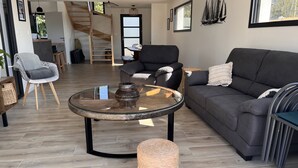 A modern living room with a glass coffee table, a grey sofa, a wicker ottoman, and a staircase leading to an upper level. - Maison de Vacances à 200 m de L'aber Benoît (Landéda)