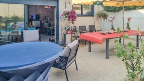 An outdoor patio with a blue table, a red table, and a yellow umbrella. There are chairs and potted plants around.