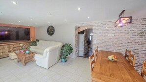 A living room with a white sofa, a wooden coffee table, and a dining table with chairs. There is a TV, a clock, and a potted plant.