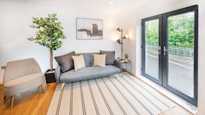 Ein modernes Wohnzimmer mit grauem Sofa, gestreiftem Teppich und einem großen Fenster mit Blick ins Grüne.