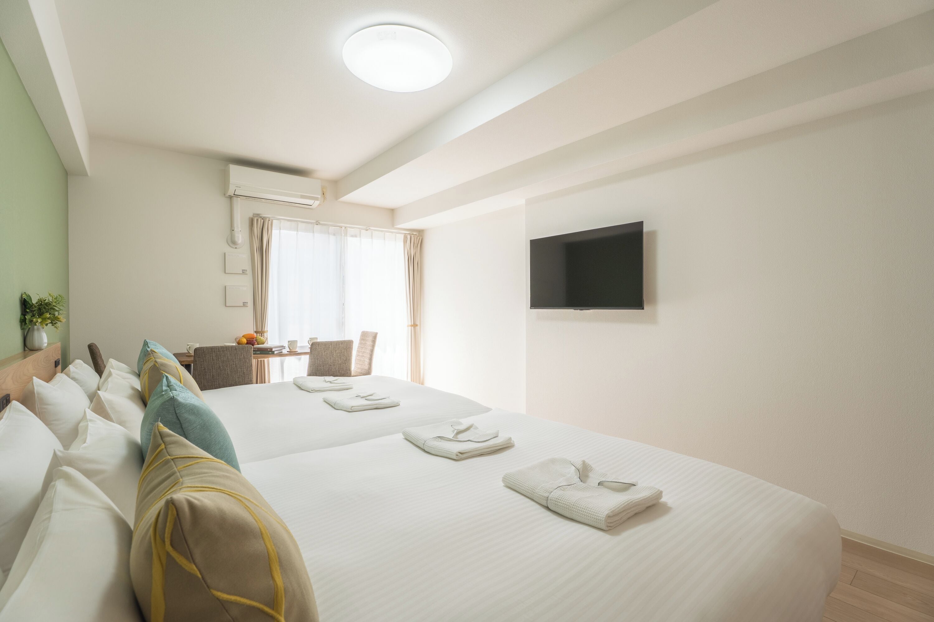 A hotel room with two beds, a flat-screen TV mounted on the wall, a dining area with a table and chairs, and a window with curtains.
