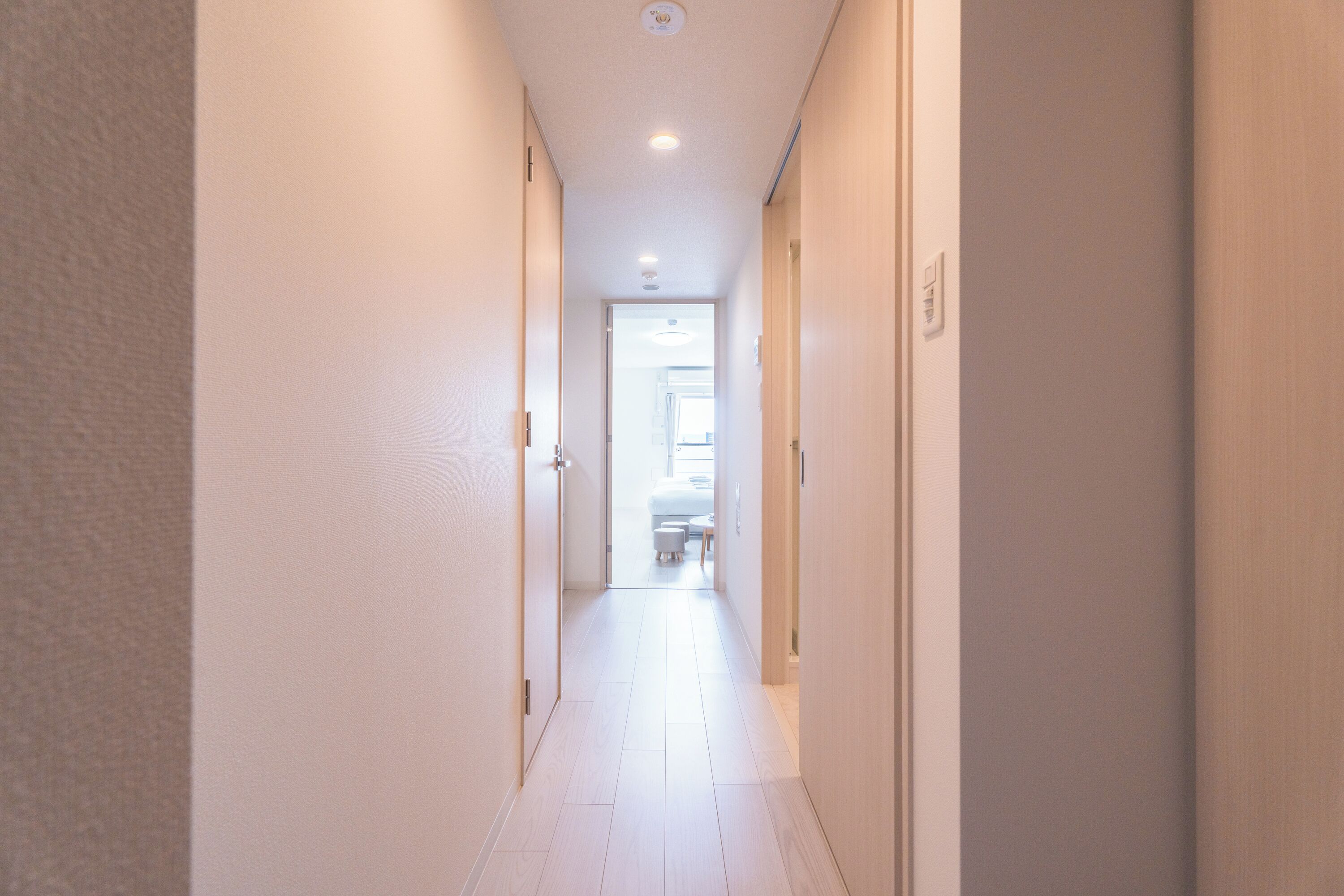 A narrow, well-lit hallway with a door at the end leading to a room with a bed and a window.
