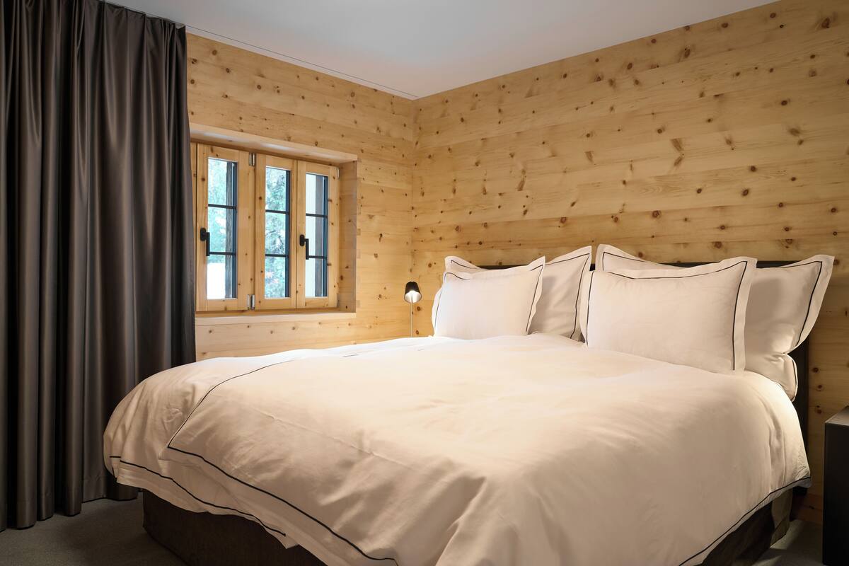 Un dormitorio con una cama grande, paredes de madera, un ventanal con cortinas y una mesita de noche con una lámpara.