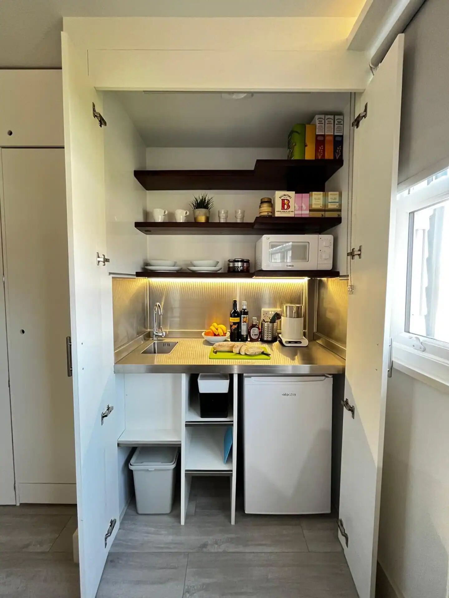 Una cocina compacta con microondas, lavabo y refrigerador pequeño. Hay repisas sobre la encimera con distintos utensilios de cocina y una ventana a la derecha.