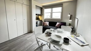 A compact kitchen with white cabinets, a dining table with chairs, a small sofa with cushions, and a window with natural light. - Apt. 12 - Luxury Living Chic Studio Steps from Beach (Brighton and Hove)