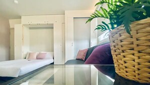 A modern bedroom with a glass table, a bed with pink pillows, a purple sofa, and a woven basket with a plant. - Apt. 12 - Luxury Living Chic Studio Steps from Beach (Brighton and Hove)