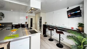 A modern kitchen with a Bosch dishwasher, a glass island counter, bar stools, and a mounted TV displaying a menu. - Apt. 12 - Luxury Living Chic Studio Steps from Beach (Brighton and Hove)
