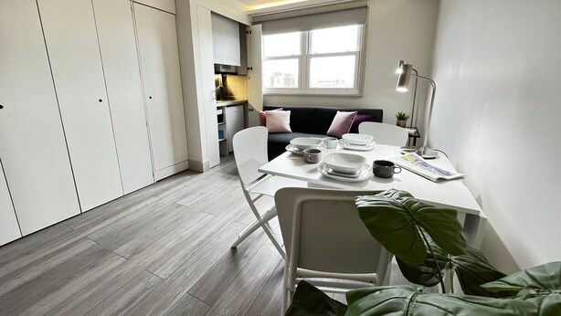 A compact kitchen with white cabinets, a dining table with chairs, a window, and a plant. - Apt. 12 - Luxury Living Chic Studio Steps from Beach (Brighton and Hove)