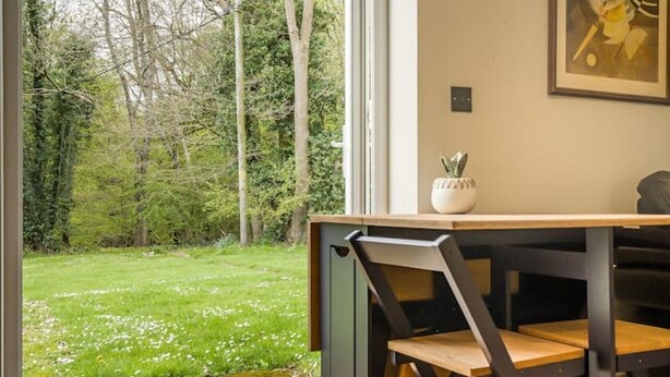 A room with a view of a grassy garden and trees. There is a table with chairs and a small potted plant on the windowsill. - Country cottage (Horsham)
