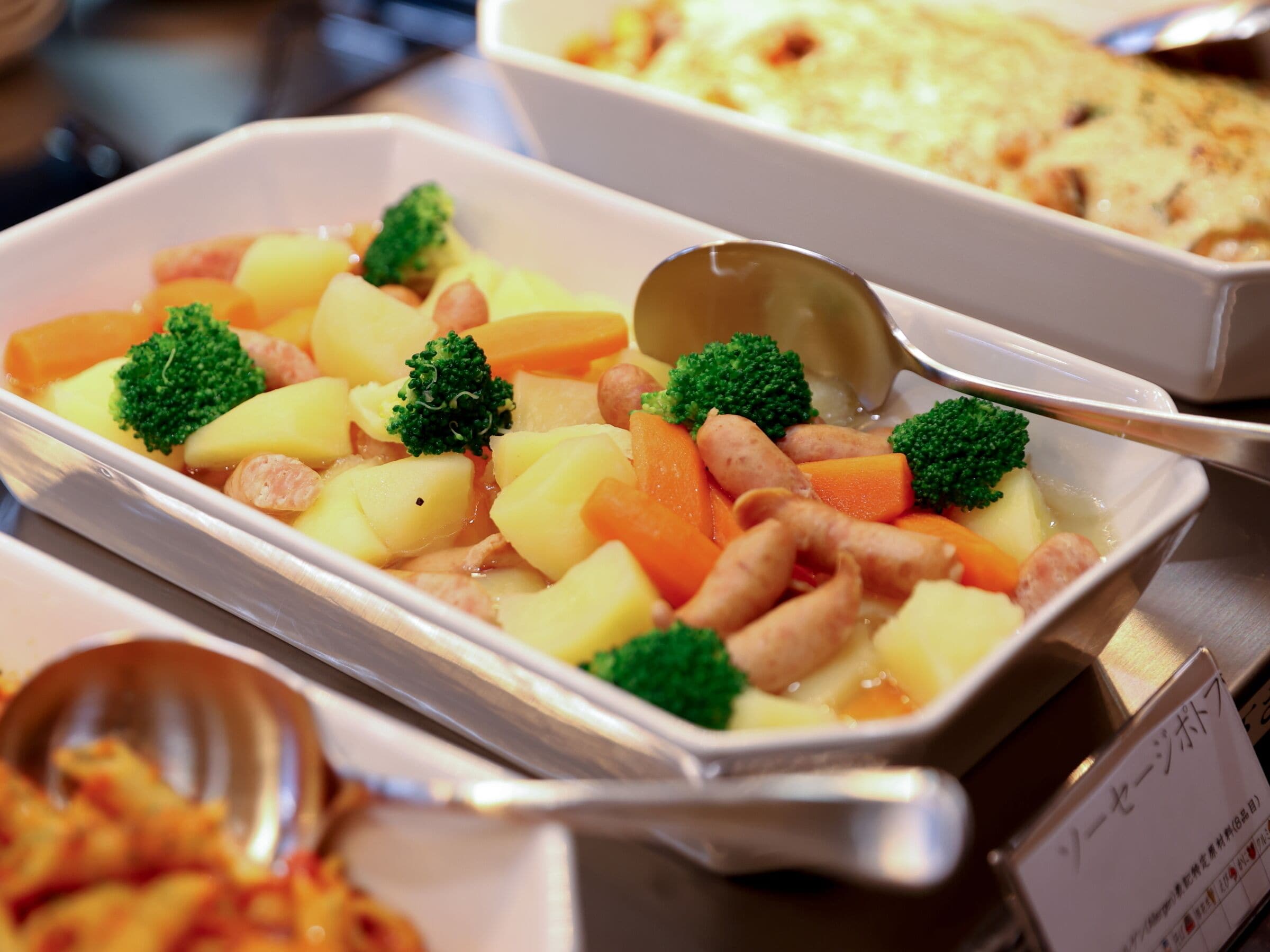 A buffet with various dishes including potatoes, carrots, broccoli, and sausages.