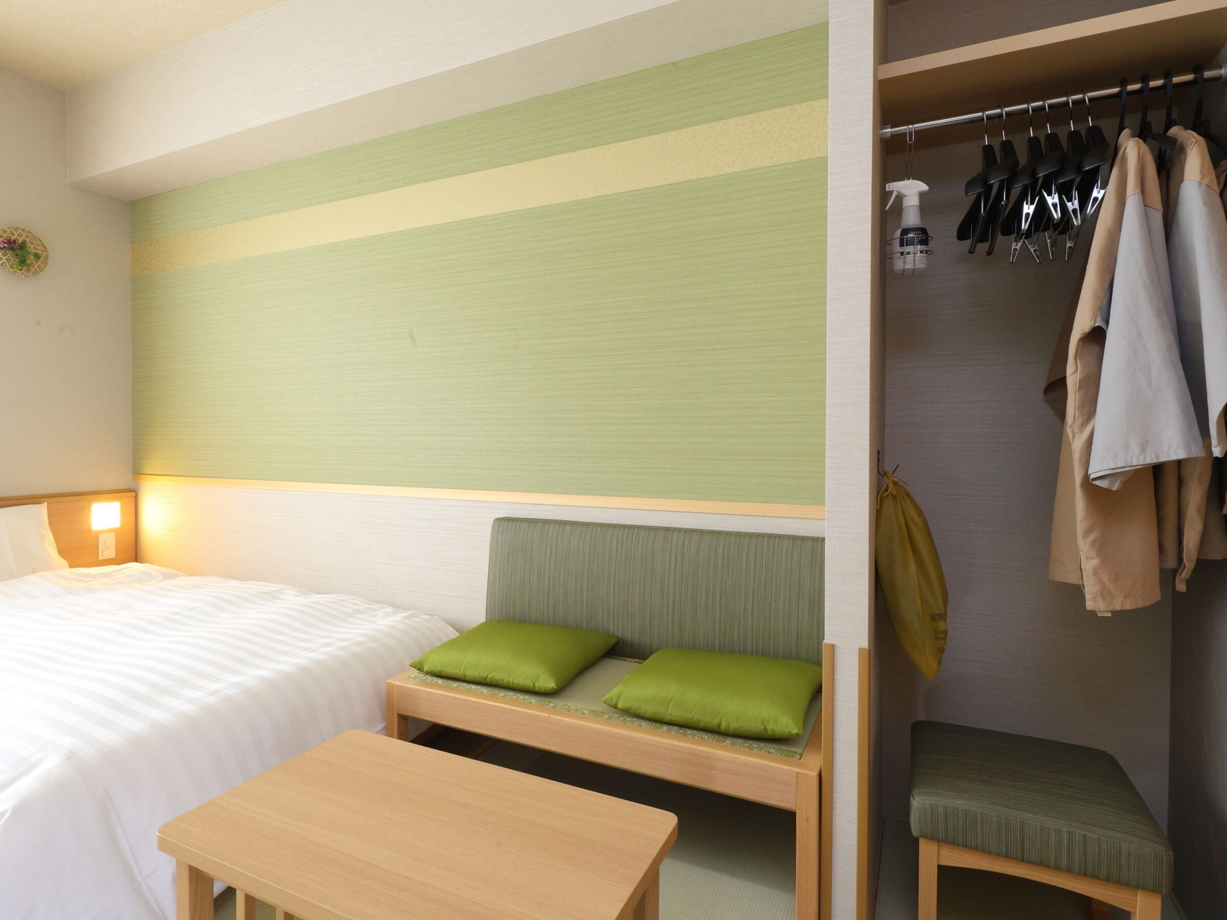 A hotel room with a bed, a small table, a bench with green cushions, and a clothing rack with hanging clothes.