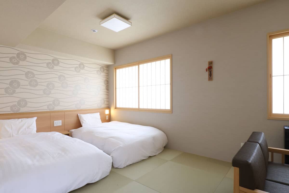 Een hotelkamer met twee bedden, een klein bureau, een stoel, een raam met zonwering en een decoratief muurpatroon.