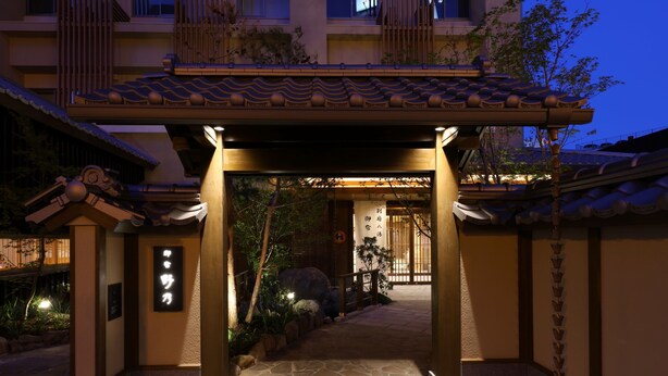 Une entrée de bâtiment japonais traditionnel, avec un toit recouvert de tuiles, un chemin de pierre et une enseigne ornée de caractères japonais.