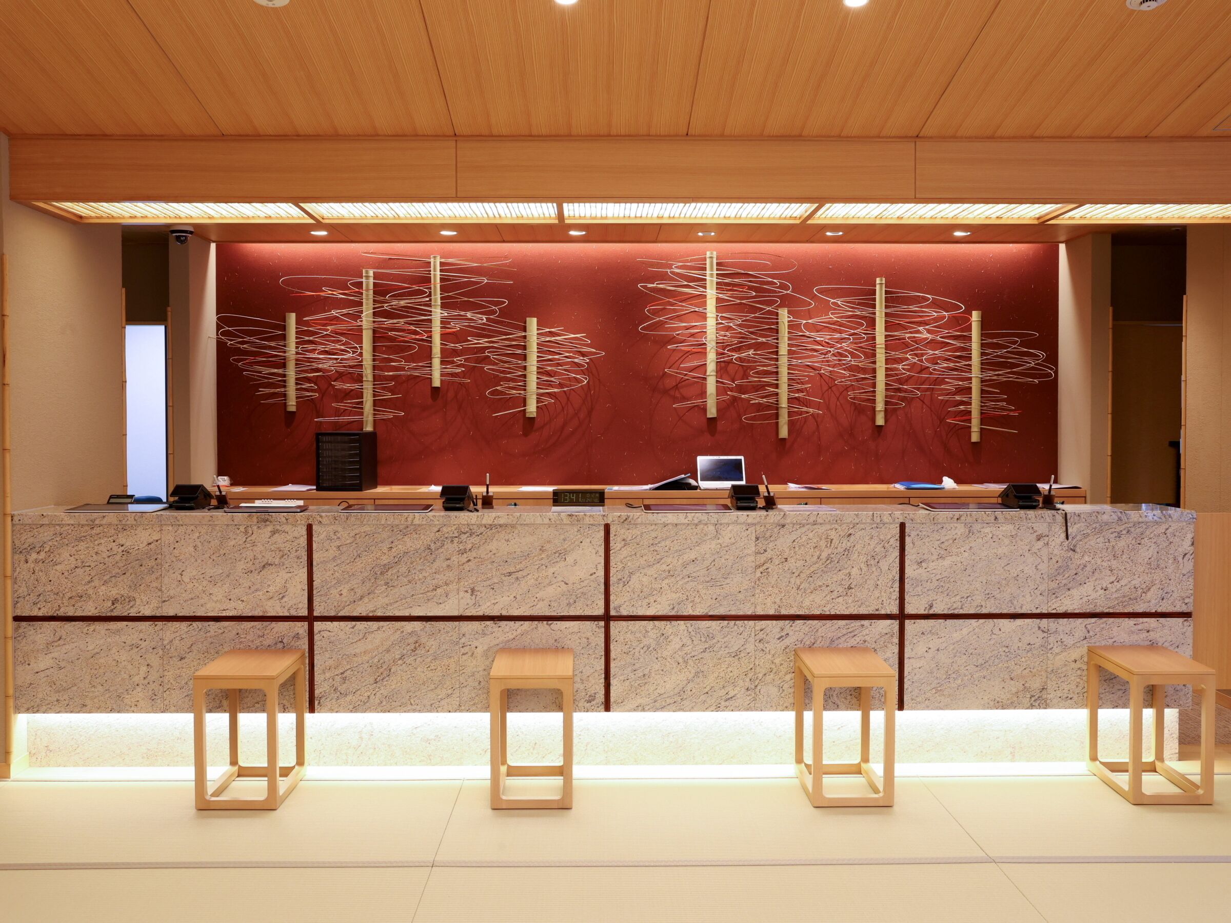 A reception desk with a red abstract wall art, a digital clock, and three wooden stools.