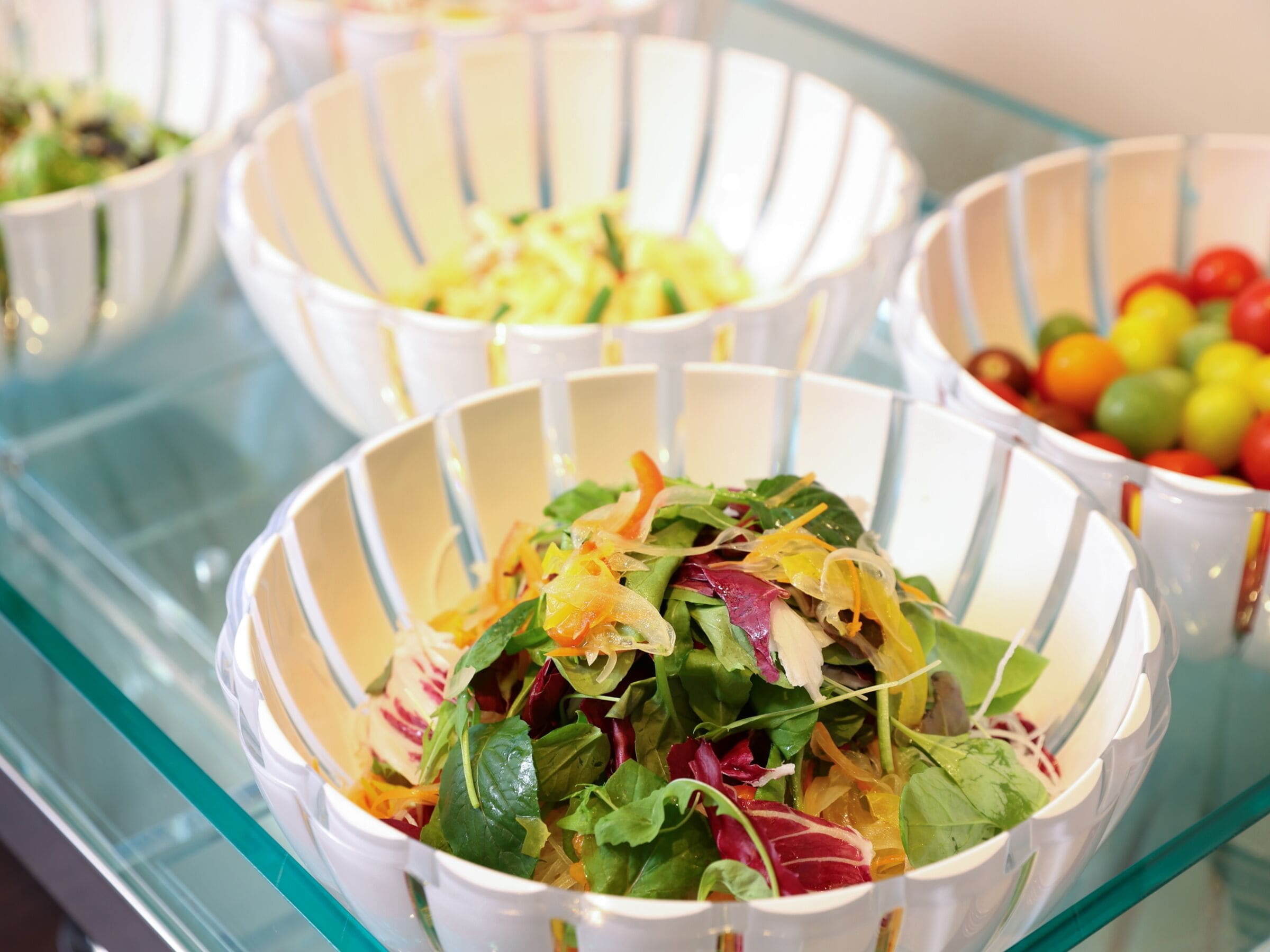 A buffet with various dishes, including salads and fruits, served in white bowls on a glass table.