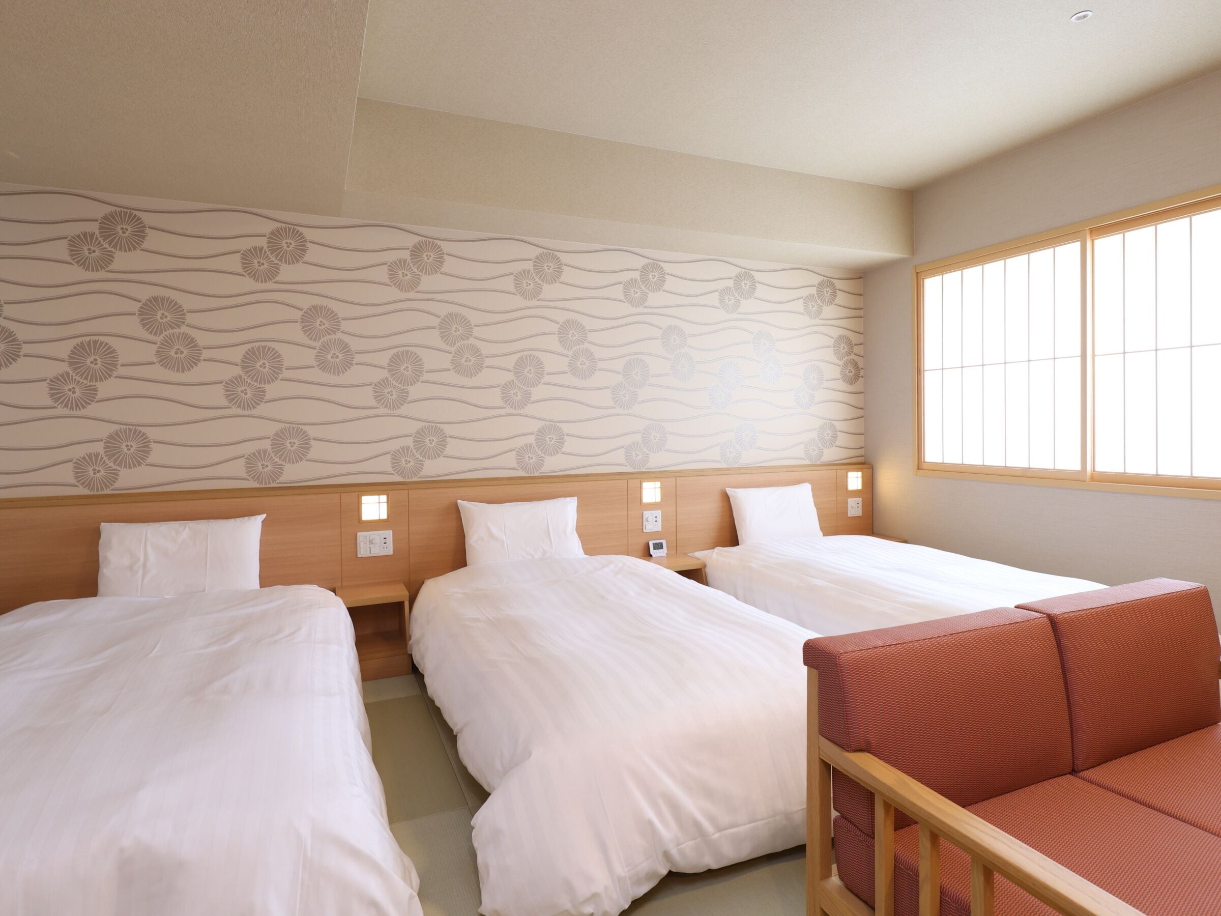 Habitación de hotel con tres camas, cabecera de madera, sofá rojo y pared decorativa con estampado floral.