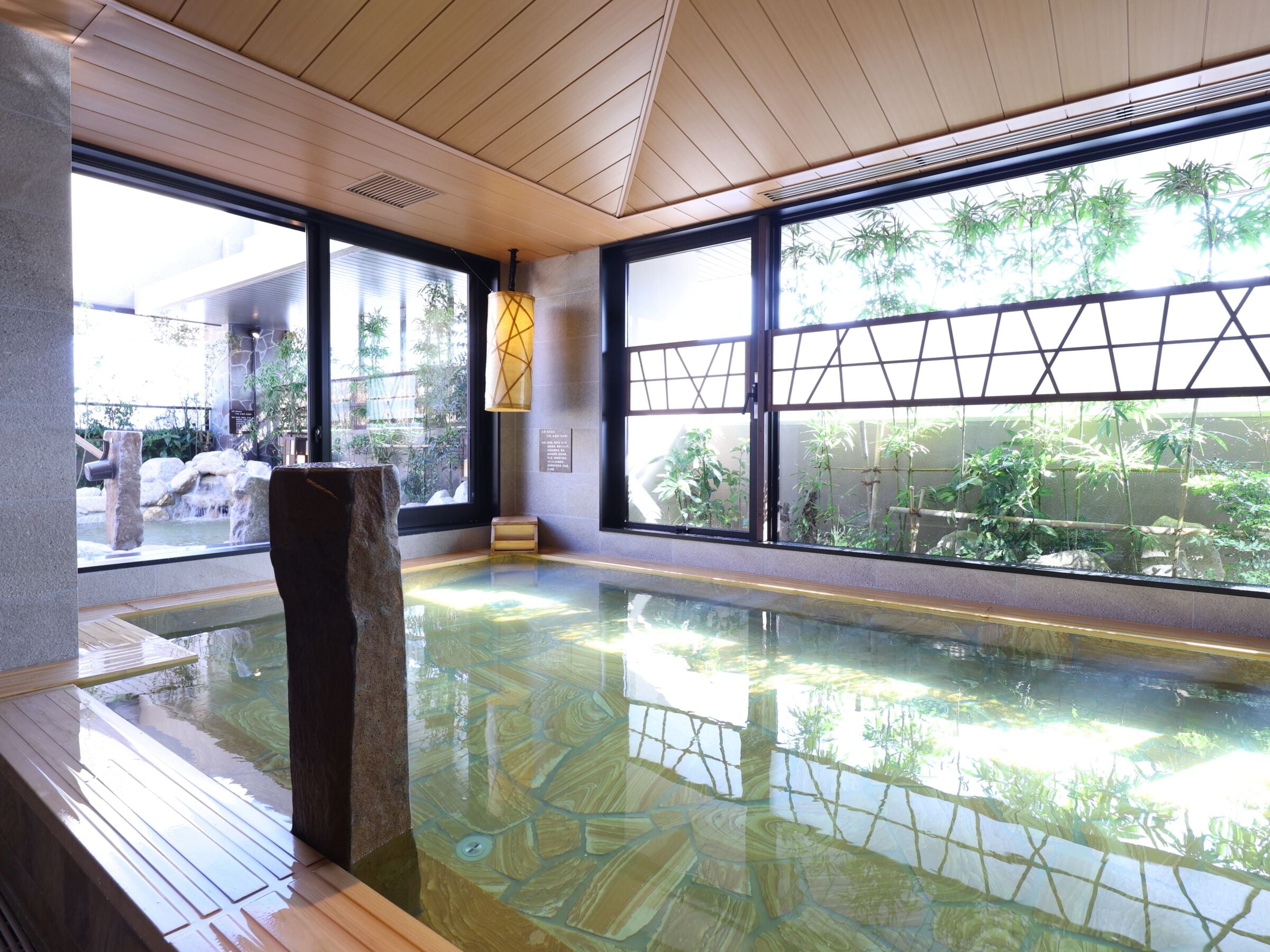 A traditional Japanese onsen with a stone bath, wooden flooring, and large windows offering views of bamboo and greenery.