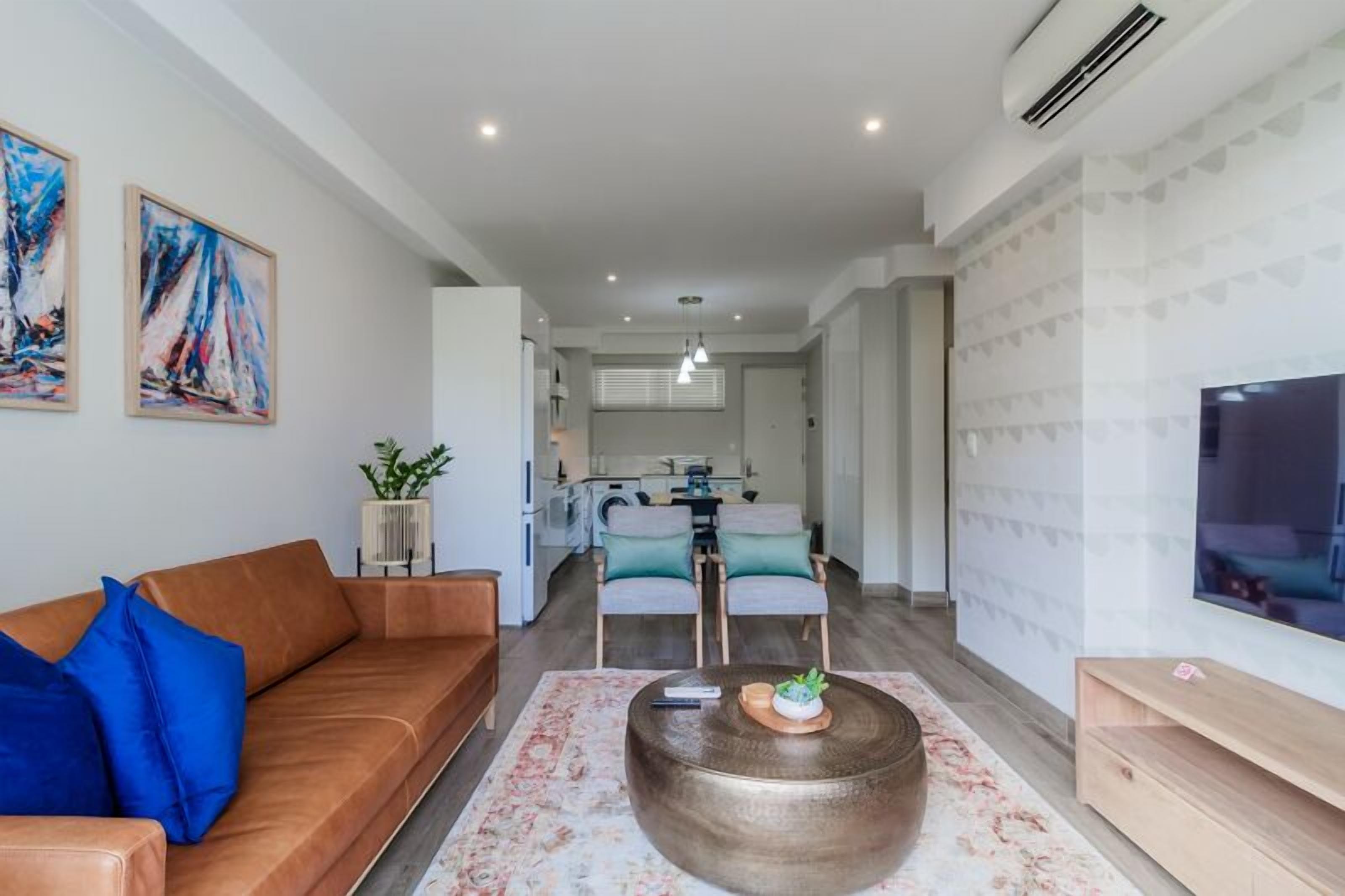 Una sala moderna con un sofá, una mesa de centro y un televisor. Se ve una cocina al fondo con un comedor.