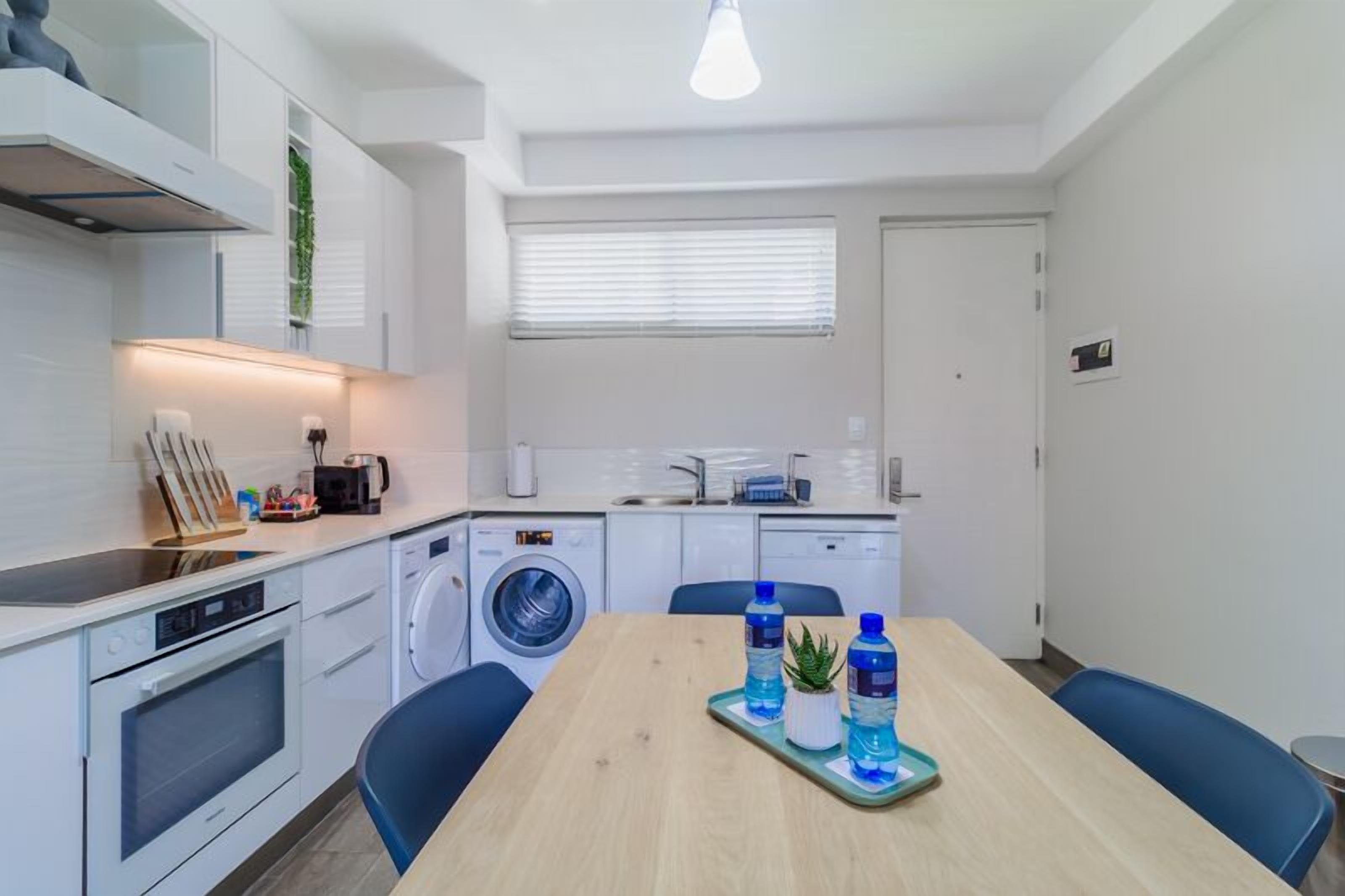 Una cocina compacta con lavadora, cafetera y mesa de comedor con sillas.