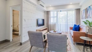 Una sala moderna con televisor de pantalla plana, sillones tapizados grises, una mesa de centro redonda, un sofá con cojines azules y una mesita auxiliar.