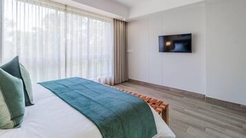 Una habitación de hotel moderna con una cama grande, un televisor montado en la pared y una ventana con cortinas translúcidas.