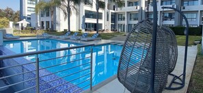 Una piscina con sillas de descanso y una sombrilla, rodeada de edificios modernos y vegetación.