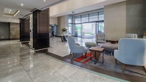 Un vestíbulo amplio con diseño moderno, que cuenta con una zona de estar con sillas y una pequeña mesa, un mostrador de recepción y grandes ventanales con vistas al exterior.