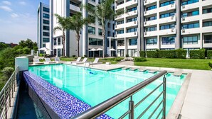 Una piscina con borde de azulejos, rodeada de sillones y sombrillas, con un edificio de varios pisos al fondo.