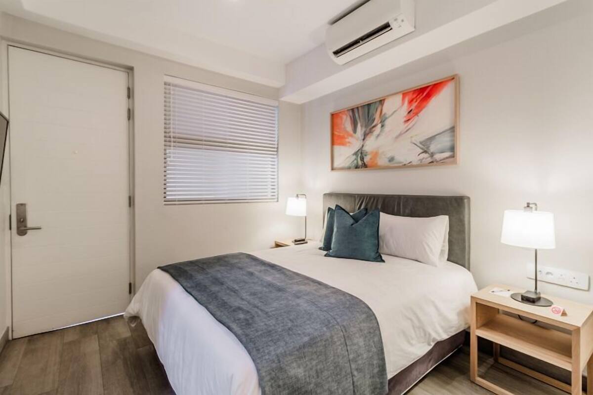 Habitación de hotel con cama, mesitas de noche, ventana con persianas, unidad de aire acondicionado y un cuadro en la pared.