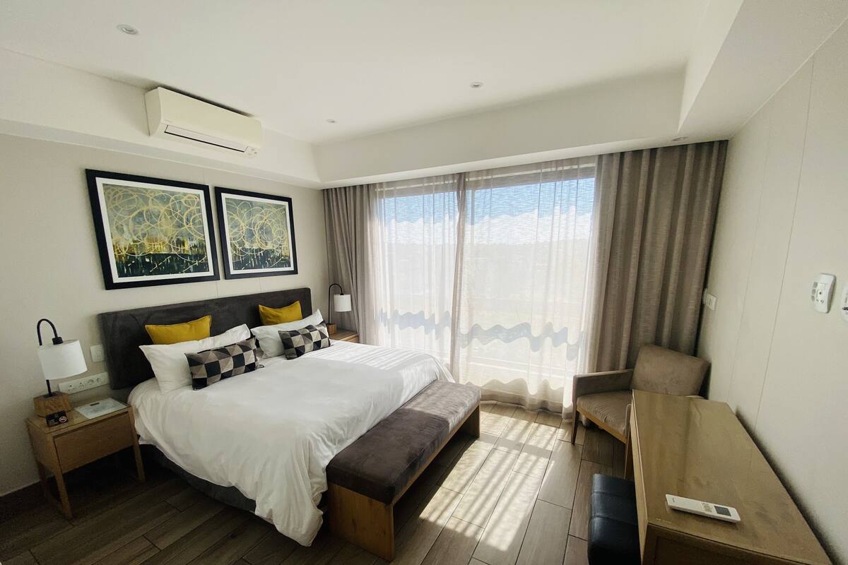 Habitación de hotel con cama, mesitas de noche, escritorio y una silla. Hay dos cuadros en la pared, una ventana con cortinas y un equipo de aire acondicionado en el techo.