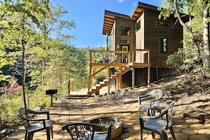 A wooden cabin with a stone patio, outdoor seating, and a staircase leading to a deck.