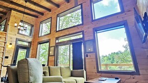 Interior acogedor de una cabaña con techo de madera, amplias ventanas, ventilador de techo y un sofá.