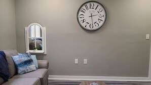 A living room with a gray wall, a large clock, a window with blinds, a gray sofa with blue cushions, and a wooden coffee table. - Cozy and Comfortable 1 bedroom (Belgium)