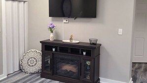 A living room with a flat-screen TV mounted on the wall, a dark wood entertainment center with a fireplace, and a patterned rug. - Cozy and Comfortable 1 bedroom (Belgium)