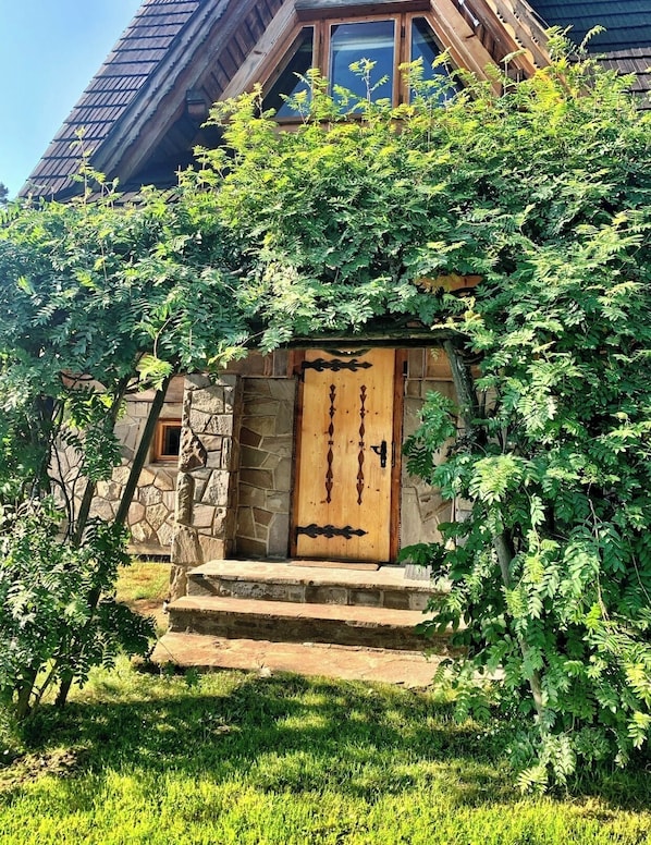 A stone house with a wooden door, surrounded by lush greenery.