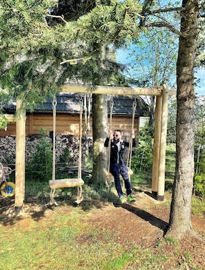 En gungställning i trä, placerad i ett skogsområde, med en byggnad synlig i bakgrunden.