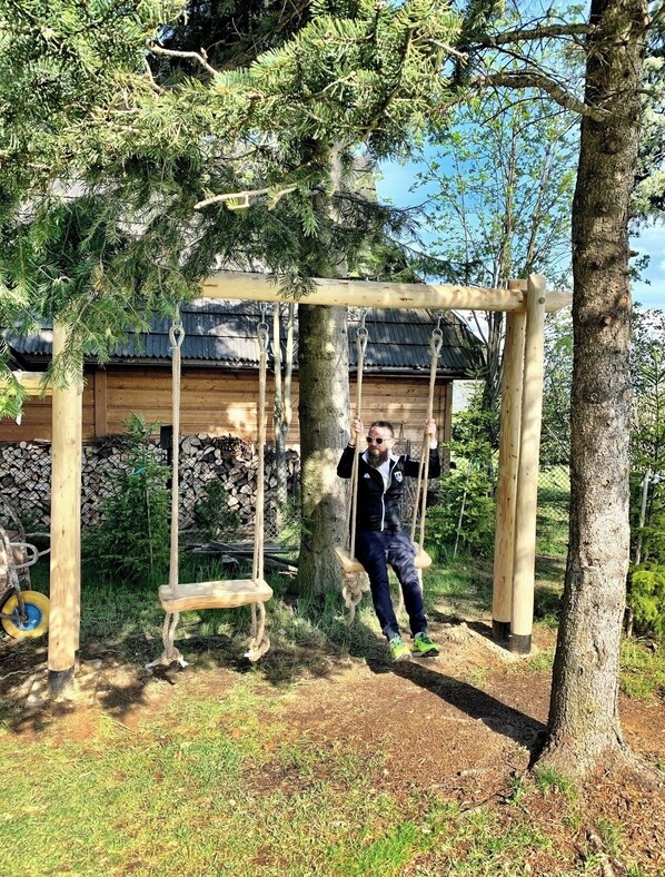 A wooden swing set in a wooded area with a building in the background. - Mountain Cozy House in Tatras With Stunning View (Poronin)