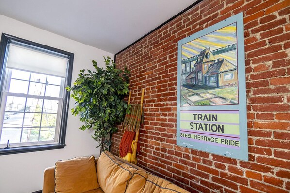 Ein Zimmer mit Backsteinwand, einem gerahmten Poster einer Bahnhaltestelle, einem Fenster und einer Couch.