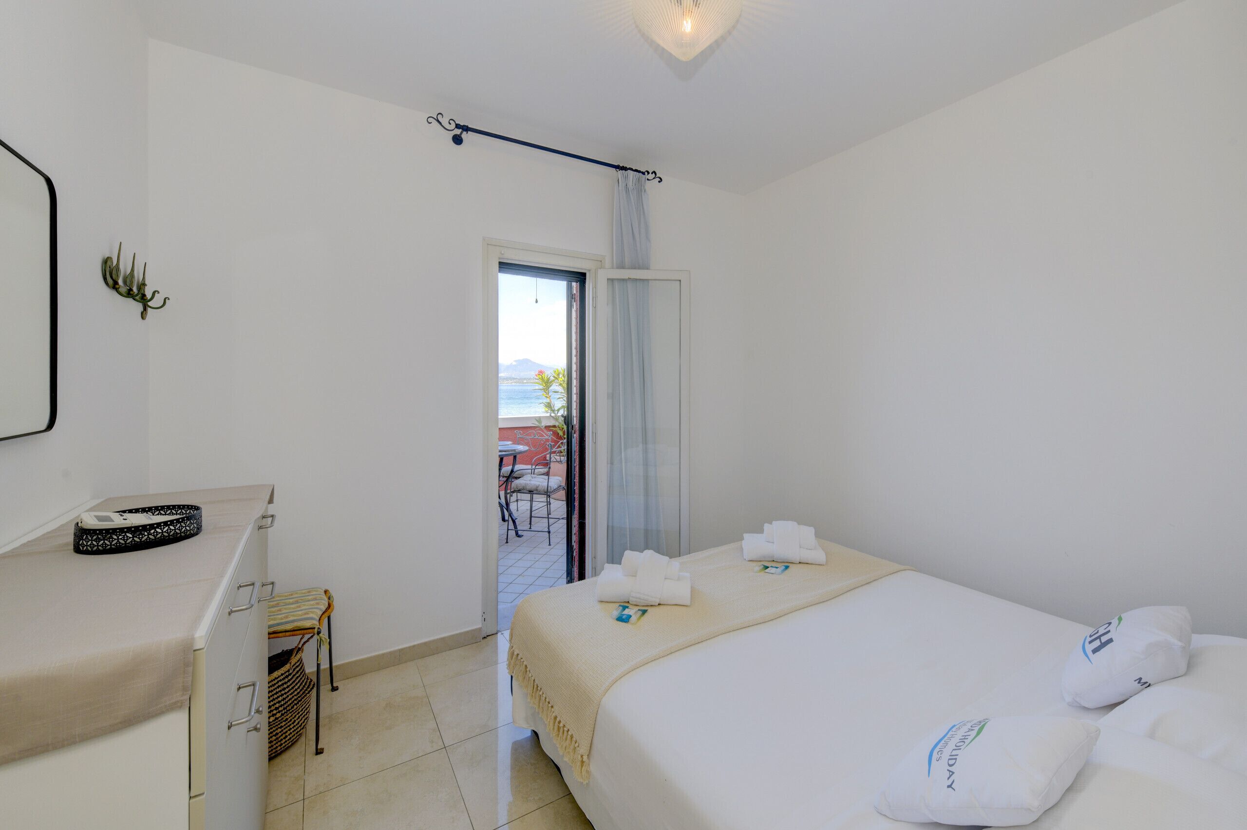 Une chambre d’hôtel avec un lit, un bureau, une chaise et une vue sur la mer, visible par une porte ouverte.
