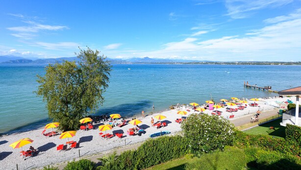 Ein Strand mit Sonnenschirmen und Liegestühlen, eine Anlegestelle und ein klarer Blick auf das Wasser und die dahinterliegenden Berge.