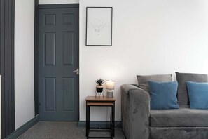 A room with a grey door, a framed picture on the wall, a small table with a lamp, and a grey sofa with blue cushions.