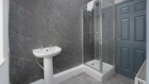 Une salle de bain avec un mur en marbre, un lavabo sur pied blanc, une cabine de douche en verre et une porte grise.