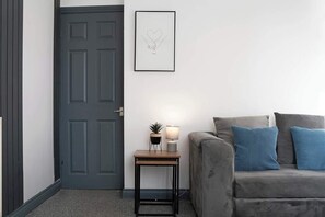 A room with a grey door, a small table with a lamp and a plant, and a grey sofa with blue cushions.